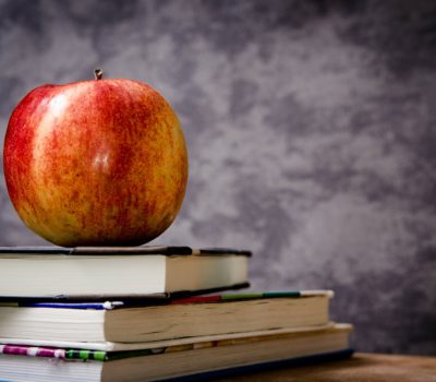An apple sits on a stack of school books in front of a a mottled grey backdrop