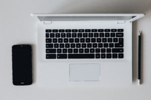 A black smartphone sits on a table next to an open white laptop and a black pencil