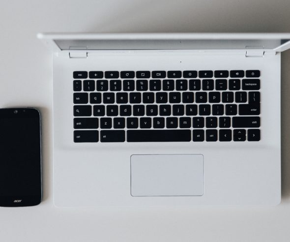 A black smartphone sits on a table next to an open white laptop and a black pencil